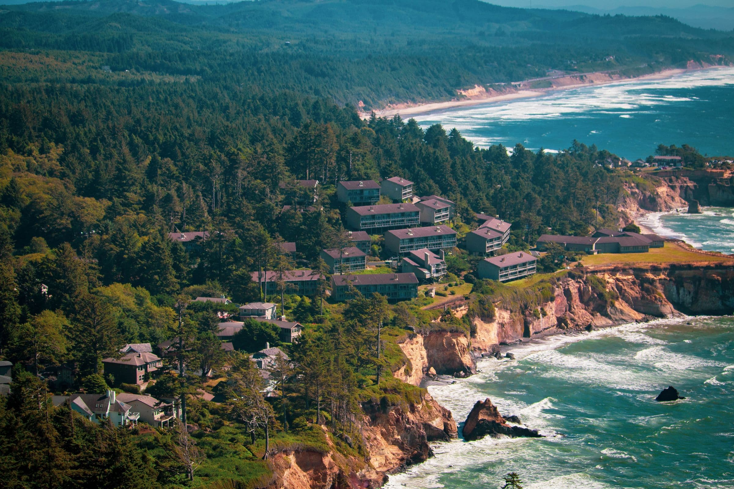 A forested headland and scattered buildings above a long coastline, seen from a distance.